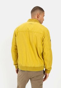 Gelbe Bomberjacke mit geripptem Kragen und Bündchen. Verfügt über sichtbare Nähte, ein Logo auf dem Rücken und einen leicht strukturierten Stoff.