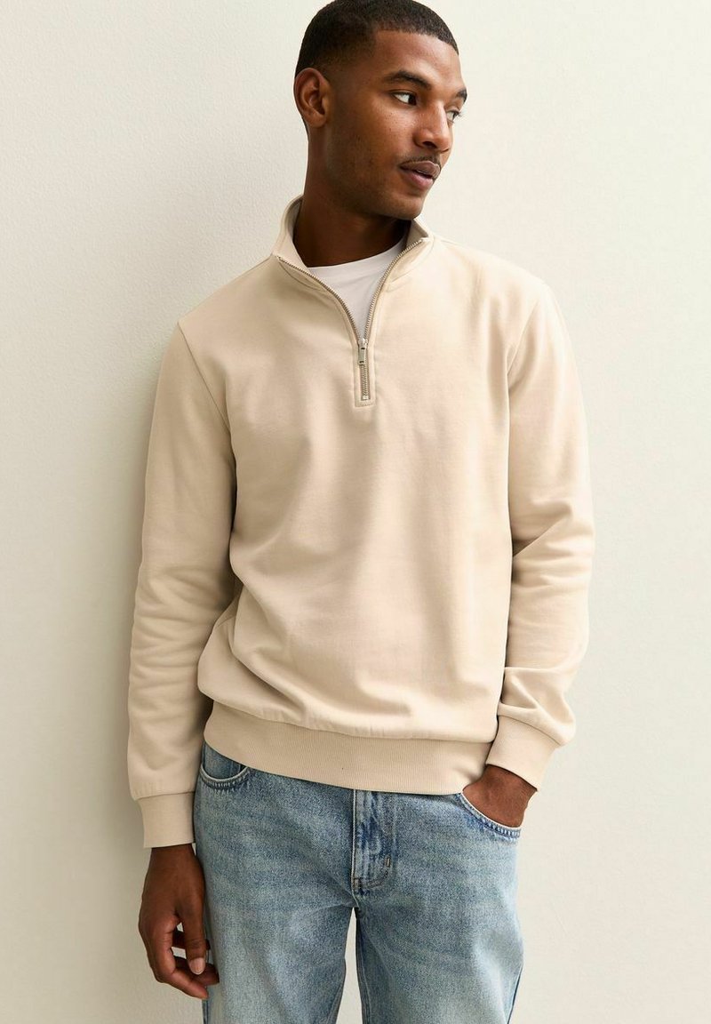 Sudadera beige con cremallera al cuarto, puños y dobladillo acanalados, llevada sobre una camiseta blanca, combinada con vaqueros azul desgastado. Textura suave, diseño casual.