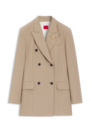 Blazer beige à double boutonnage avec boutons noirs, revers crantés, poches à rabat et broche dorée en forme de fleur sur le revers gauche.