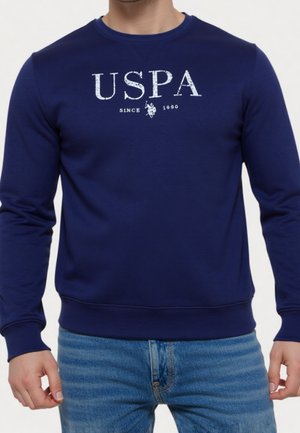Homme portant un sweat-shirt bleu marine avec « USPA Since 1890 » et un logo de joueur de polo imprimé en blanc sur la poitrine.