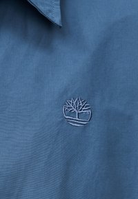 Μπλε πουκάμισο από ύφασμα με μυτερό γιακά και μικρό κεντημένο λογότυπο δέντρου κοντά στο κέντρο.