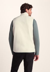 White fleece vest with high collar and textured fabric, worn over a teal long-sleeve shirt. Back view shows simple design and no visible hardware.