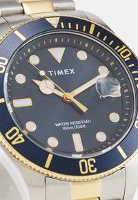 Montre Timex avec une lunette bleue foncée, des accents dorés et un cadran noir avec de grands index horaires. Résistante à l'eau jusqu'à 100 m. Bracelet en acier inoxydable.