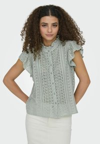 Lichtgroene kantblouse met gerimpelde mouwen, knoopsluiting en gedetailleerde uitsnijdingen door de stof. Gladde textuur.