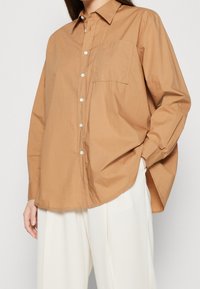 Camicia button-up in cotone marrone con colletto a punta, una tasca sul petto e maniche lunghe, abbinata a pantaloni bianchi a gamba larga.
