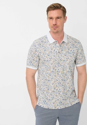 Man met kort blond haar, gekleed in een korte mouw poloshirt met een bloemmotief, witte kraag en manchetten, handen in de zakken, kijkt opzij.