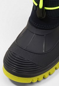 Botte noire imperméable avec semelle en caoutchouc texturé et accent jaune. Dispose d'un dessus en mesh, de lacets réglables avec un cordon de serrage, et de détails nervurés.