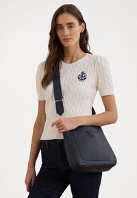 Navy Leder Schultertasche mit geprägt Logo, rechteckiger Form, verstellbarem Tragegurt und glatter Textur. Getragen mit einem cremefarbenen Strickoberteil.