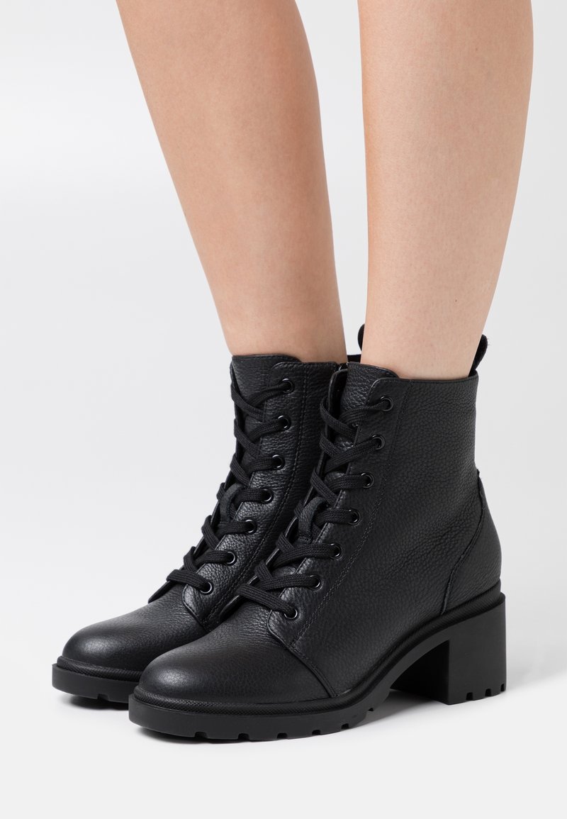 Bottes basses à lacets en cuir noir avec talons blocs épais portées sur des jambes nues sur un fond clair uni.