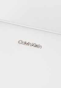 Calvin Kleinin hopea logo valkoista teksturoitua taustaa vasten, jossa on tyylikäs, minimalistinen muotoilu puhtain linjoin.
