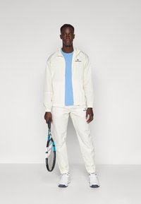 Hvitt sportsjakke med blå logo, kombinert med en lyseblå T-skjorte og hvite joggebukser. Holder en tennisracket, iført hvite joggesko.