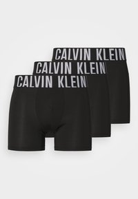 Black boxers made of stretchy fabric, featuring a wide waistband with "Calvin Klein" printed in white. Three pairs included in the set.