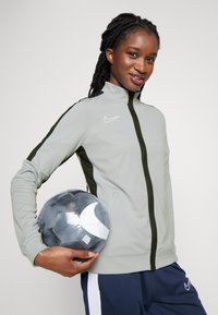 Lichtgrijze ritsjack met zwarte zijaccenten, gemaakt van gladde stof, gecombineerd met donkerblauwe broek en met een zwart-wit voetbal in de hand.