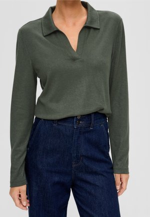 Kvinde iført en mørkegrøn langærmet polotrøje, der er stukket ned i højtaljede mørkeblå jeans med to synlige knapper.