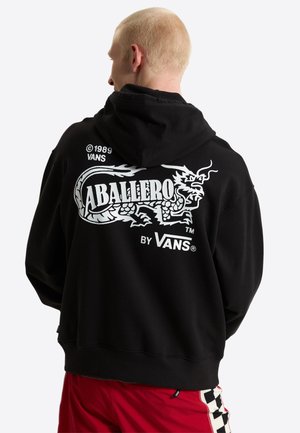Persoon met kort blond haar die een zwarte hoodie draagt met een witte draakafbeelding en de tekst "Caballero by Vans" op de achterkant.