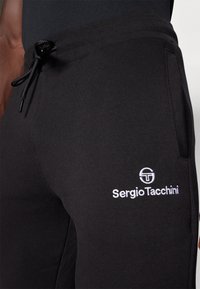 Črne bombažne trenirke z vrvico v pasu, stranskimi žepi in belim vezenim logotipom ali besedilom na spodnjem delu noge.