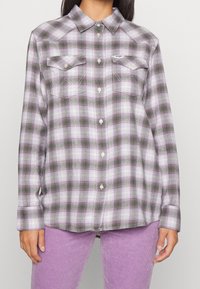 Camicia di flanella a quadri con colori viola, verde e bianco; presenta due tasche sul petto, chiusura con bottoni e colletto classico.