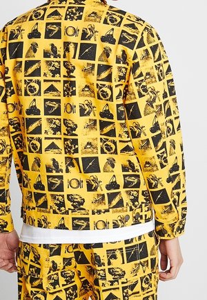 Gul jakke og bukser med sort grafisk print af forskellige abstrakte former og genstande, båret af en person vist bagfra.