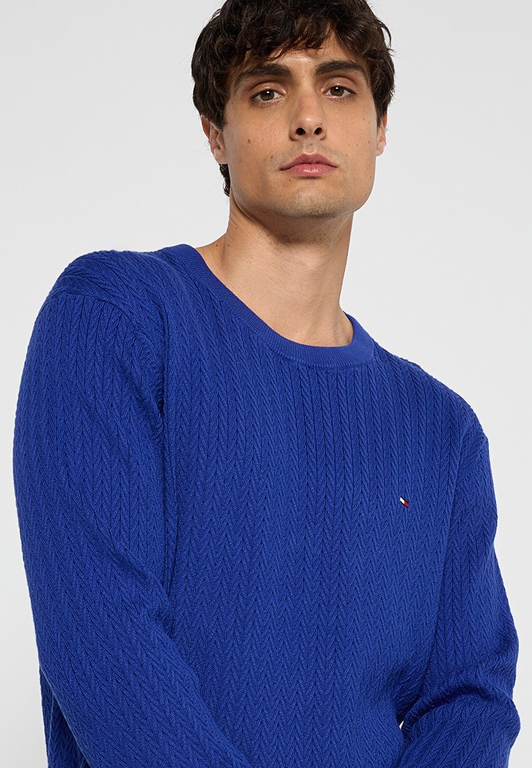 Blauwe gebreide trui met een gestructureerd herringbone patroon, ronde halslijn en een klein logo op de borst. Zacht, mediumgewicht materiaal.