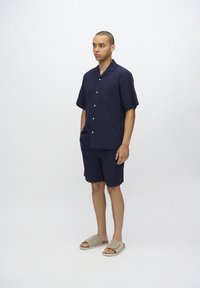 Námornícka krátkopyjový button-up košeľa s vreckom, spárovaná s ladiacimi šortkami. Model nosí béžové šľapky. Hladká, štruktúrovaná tkanina.