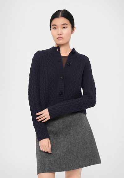 Navy blue knitted cardigan with textured cable pattern, five buttons, and ribbed cuffs, paired with a gray houndstooth skirt.