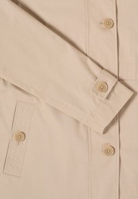 Chemise beige à manches longues avec boutons aux poignets, sur la patte de boutonnage avant, et une poche rectangulaire cousue.