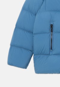 Sininen tikattu puffer-takki, jossa on hihat ja etuosa, joissa on musta pystysaumattu tasku valkoista taustaa vasten.