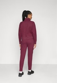 Trening maron cu guler cu fermoar, manșete cu model și buzunare cargo pe pantaloni. Stilizat cu adidași negri și șosete albe.