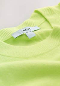 Pull à col rond côtelé vert clair avec une étiquette en tissu blanc portant la mention "NEXT" cousue à l'intérieur du col.