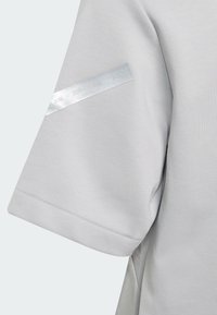 Camiseta gris claro de manga corta, con una banda plateada reflectante en la manga que muestra los años 2018 a 2021.