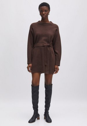 Robe tricotée marron avec de longues manches et une taille ceinturée. Texture lisse, poignets côtelés, associée à des bottes en cuir noir jusqu'aux genoux.