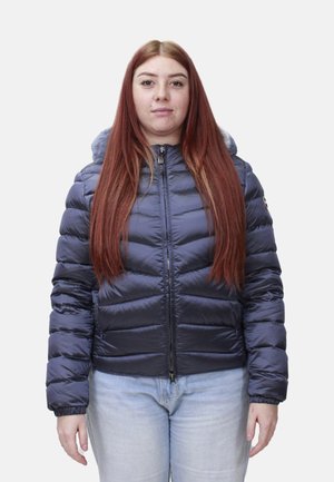 Donna con lunghi capelli rossi che indossa una giacca imbottita blu scuro con zip e jeans azzurri, in piedi su uno sfondo semplice.