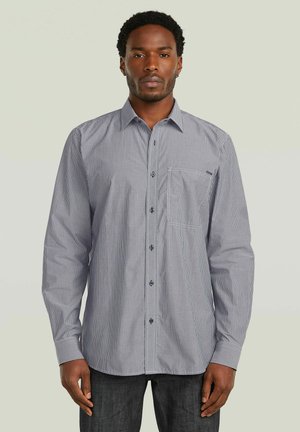 Mand iført en langærmet, sort-hvid tyndstribet skjorte med knapper og brystlomme, stående mod en ensartet lys baggrund.