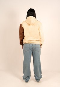 Sudadera con capucha en color crema, marrón y beige claro, con un corte relajado. Combinada con jeans de pierna ancha en azul claro que tienen bolsillos traseros.