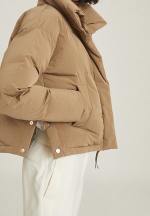 Person wearing a tan puffer jacket with hands in pockets, paired with white pants against a plain light background.