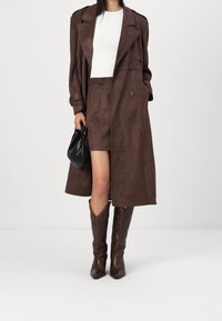 Kvinna som bär en lång brun trenchcoat, matchande kjol, vit topp, bruna cowboyskor och håller en liten svart handväska.