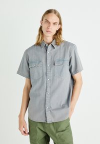 Camisa de manga corta, con botones, hecha de tela de chambray azul claro. Presenta dos bolsillos en el pecho y un cuello con botones a presión.