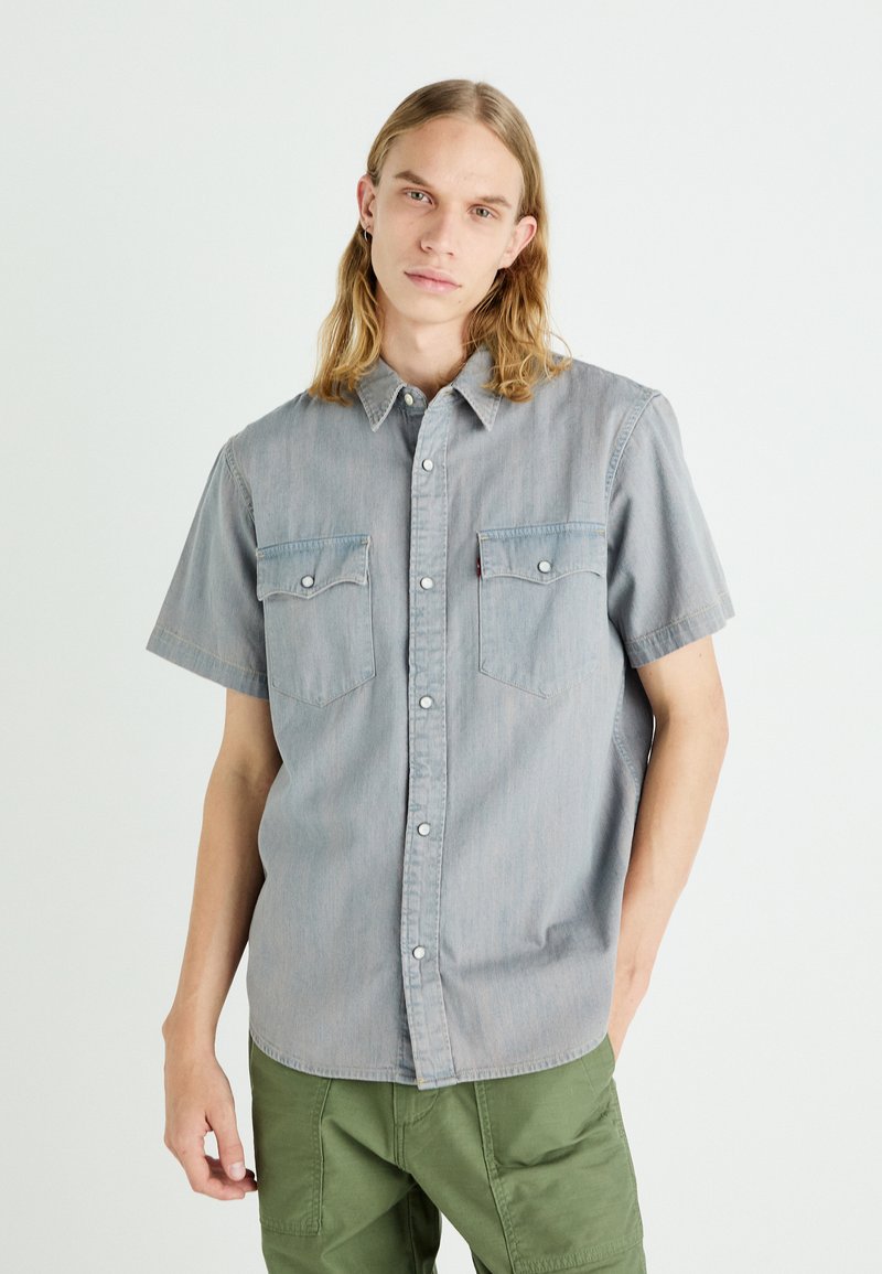 Camisa de manga corta, con botones, hecha de tela de chambray azul claro. Presenta dos bolsillos en el pecho y un cuello con botones a presión.