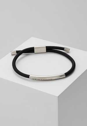 Bracelet à double cordon noir avec des accents métalliques argentés, comprenant une barre courbée gravée de "Armani Exchange,", présenté sur une surface géométrique blanche.