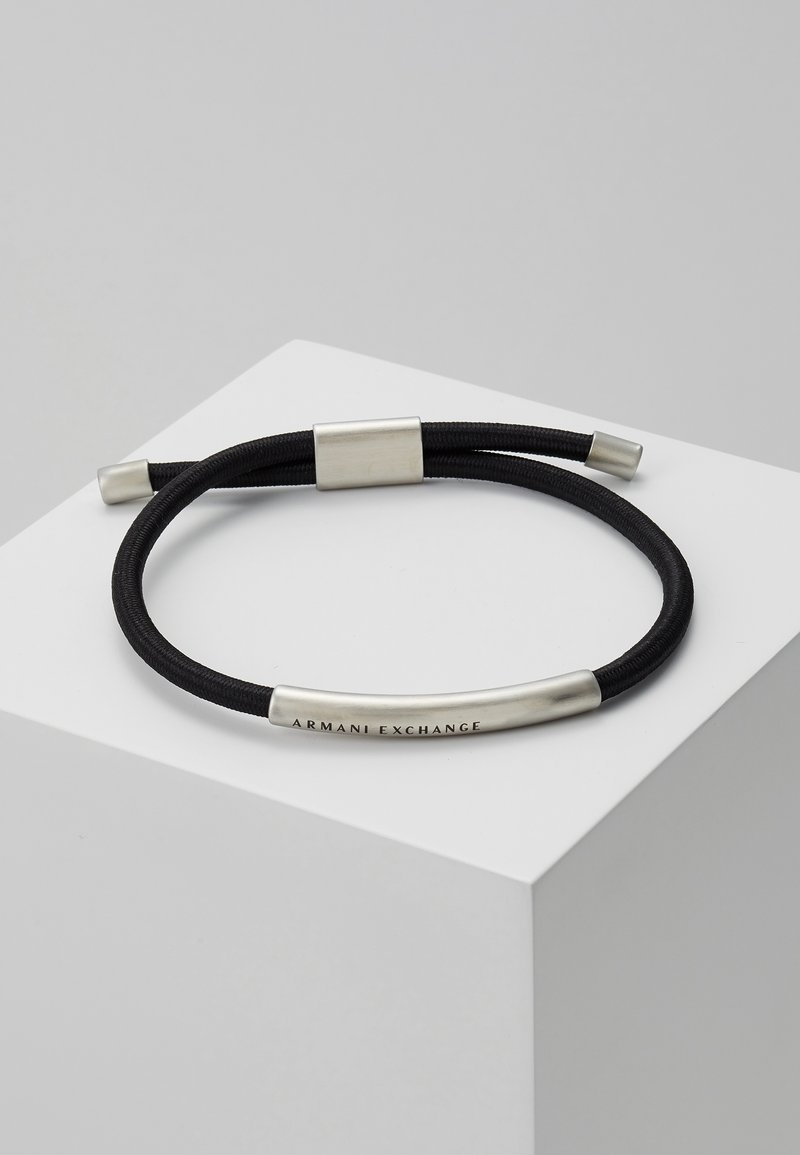 Bracelet à double cordon noir avec des accents métalliques argentés, comprenant une barre courbée gravée de "Armani Exchange,", présenté sur une surface géométrique blanche.