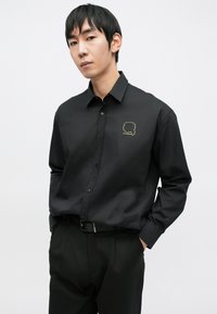 Crna košulja s gumbima izrađena od glatkog materijala, s zlatnim grafikonom na lijevoj strani prsa. Nošena s crnim hlačama i remenom za elegantan izgled.