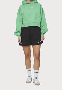 Sudadera verde corta con bolsillo frontal; pantalones cortos negros; calcetines blancos con logo negro; zapatillas gris y verde. Tela sencilla y diseño casual.