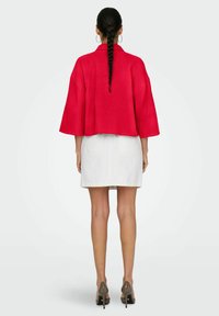 Giacca rossa tagliata con un tessuto testurizzato e maniche larghe, abbinata a una gonna di jeans bianca e scarpe con tacco beige.