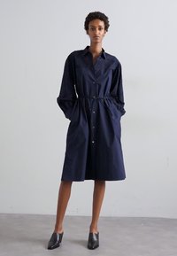 Granatowa sukienka zapinana na guziki z wcięciem w talii, długimi dzwonowymi rękawami oraz teksturowanymi akcentami na ramionach. Czarne buty dopełniają całość.