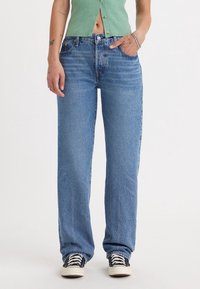 Vaqueros de mezclilla azul claro con un corte recto y ligero desgaste. Presenta cinco bolsillos y sutiles detalles de costura, combinados con zapatillas negras.