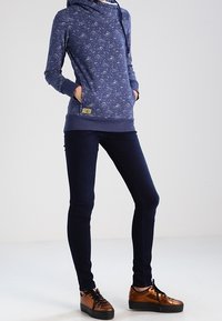 Granatowa bluza z kapturem z wzorem, z troczkiem i przednimi kieszeniami, noszona z dopasowanymi ciemnymi dżinsami oraz brązowymi tenisówkami z czarnymi podeszwami.