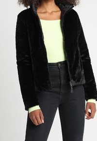 Svart fuskpälsjacka med dragkedja framtill, hög krage och uppvikta ärmar, bärs över en ljusgrön topp, ihop med svarta jeans.