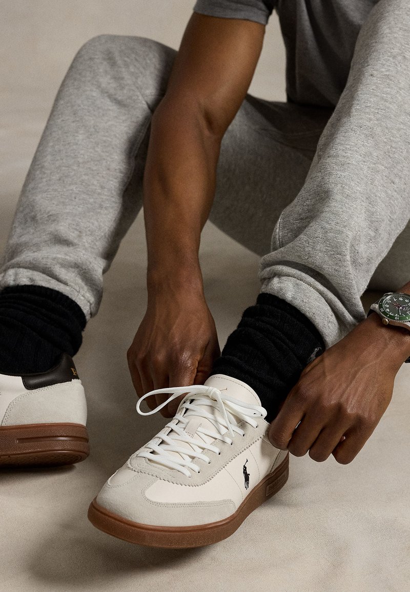 Persoon draagt grijze joggingsbroek en zwarte sokken en maakt de veters vast van witte sneakers met bruine zolen en een zwart logo aan de zijkant.