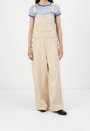 Personne portant une salopette large rayée beige et une chemise à manches courtes rayée bleue avec des chaussures noires, debout sur un fond blanc.