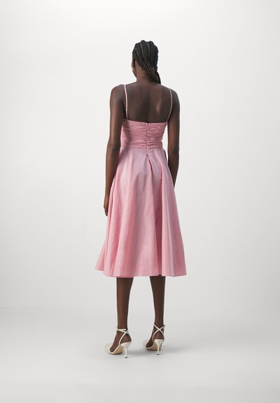 Roze midi-jurk met een aansluitende bodice en een uitlopende rok. Voorzien van dunne bandjes en een gerimpelde achterkant. Gecombineerd met gespsandalen met hoge hakken.